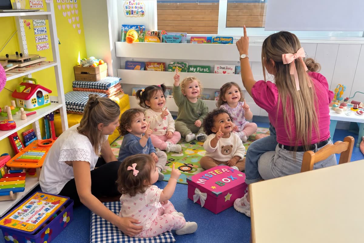 Miss Kelly leading a circle of toddlers who point up and smile, with a pink mystery box in the middle